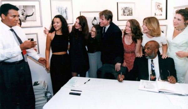 Howard Bingham getting ready to sign his book of photos of Ali at his Govinda Gallery exhibition while Ali charms the Govinda Girls with some magic. © Govinda Gallery Archive